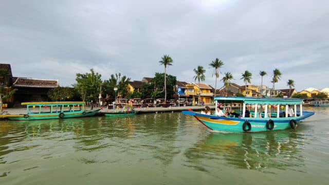 Colorful boats on a river in Hoi An with people onboard, palm trees, and yellow buildings in the background.