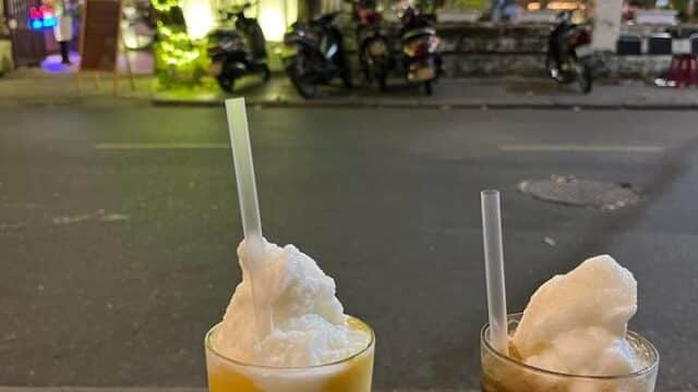 Two frothy iced drinks on a table outside, with Hoi An’s lively, decorated street glowing in the background at night.
