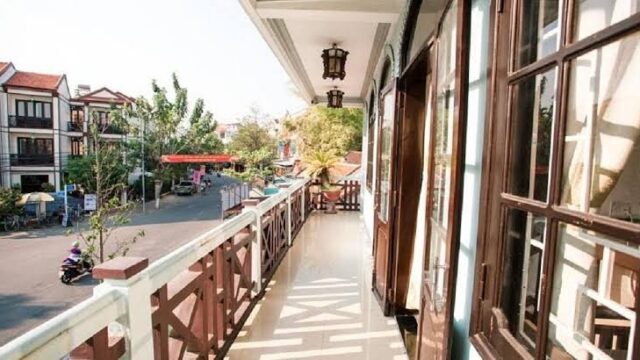A sunny balcony at Hoi Pho Hotel overlooks a quiet street lined with shops, trees, people, and motorcycles.