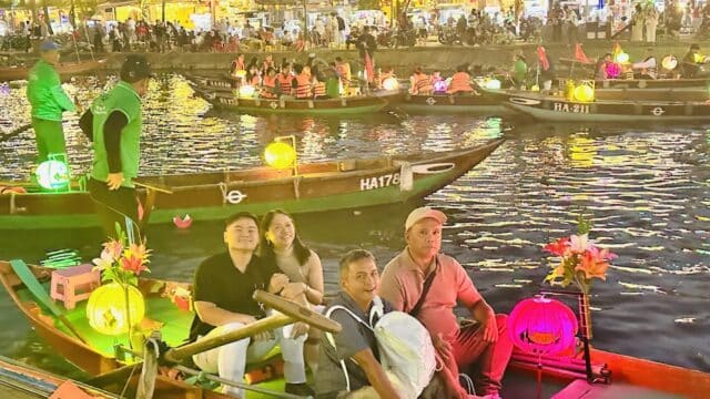 Four people sit in a lantern-lit boat on a river at night, enjoying Hoian tours amid colorful boats and lively crowds.