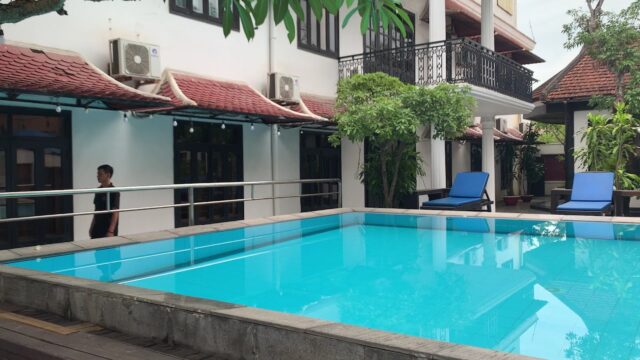 A courtyard swimming pool at Hoian Nostalgia Hotel, with blue lounge chairs and a person walking nearby.