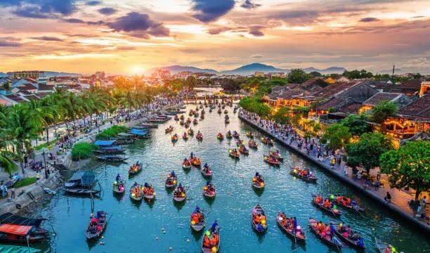 Colorful boats with people on a river at sunset, lined by palm trees and busy streets capture authentic Hoian Vibes in this vibrant town.