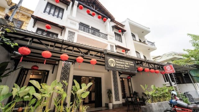 White building with red lanterns, Old Town Hotel sign, Hoianese charm, lush plants, and a motorbike parked outside create a quiet scene.