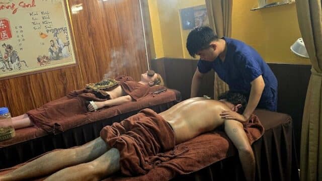 Two people receive massages in a dimly lit spa room; one therapist works while steam rises from a back treatment.