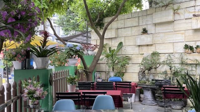 Outdoor café in Hoi An with blue chairs, black tables, lush plants, and flowers beneath trees and a striped awning.