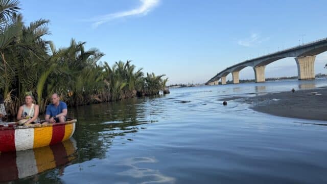 Two people sit in a colorful Hùng Hên Boat Tours vessel on a river, with palm trees and a large bridge in the background.