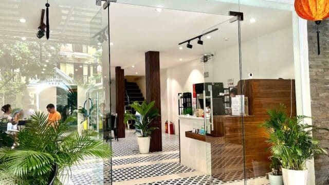 Modern café interior inside a local budget hotel, featuring glass doors, patterned tile floor, potted plants, and a coffee counter.