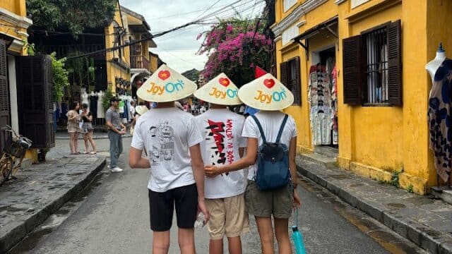 Three people wearing conical hats and shorts stand on a street with yellow buildings in an old town.