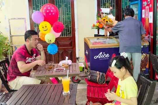 A man and a girl sit at a café table in Hoi An with drinks, balloons, and a dog; two people stand at the counter.