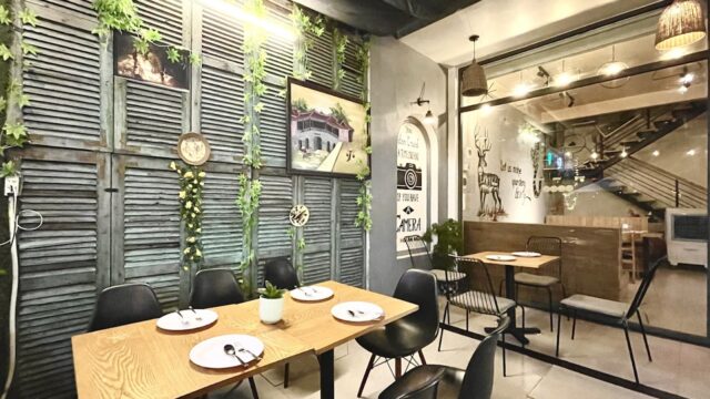A cozy Indian restaurant with wooden tables, black chairs, wall art, and hanging greenery for decoration.