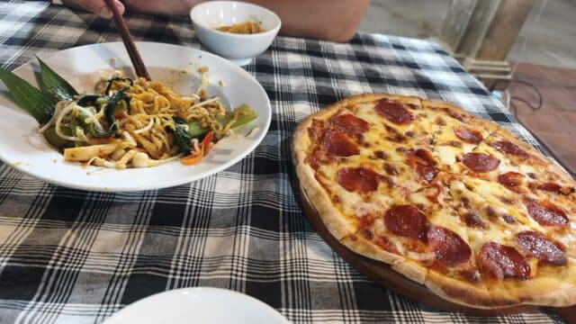 A person eats noodles with chopsticks next to a pepperoni pizza at Kitchen Town Restaurant on a checkered tablecloth.