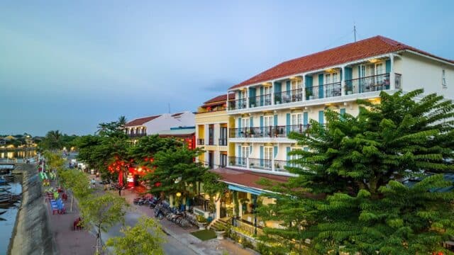 A riverside street lined with trees and colorful buildings at dusk, featuring a boutique hotel and bikes parked along the sidewalk.