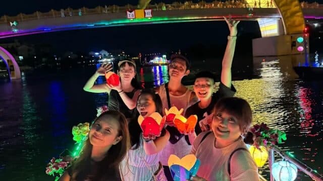 A group of friends enjoys a Lantern Boat Tour at night, holding lanterns with a lit-up bridge glowing in the background.