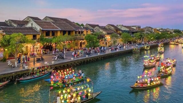 Colorful lantern boats float on a river at sunset in lively, historic Hoi An, with people along the charming waterfront.