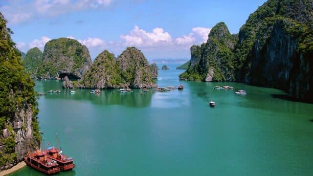Emerald green bay with limestone islands, boats, and lush cliffs under a bright blue sky near Hoi An.