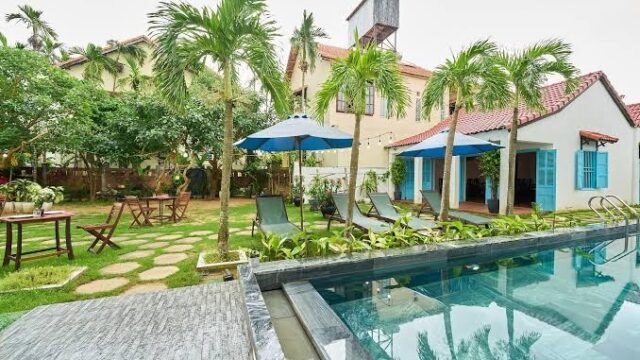 A swimming pool with lounge chairs, umbrellas, and palm trees in a lush garden at Legend Hotel’s tropical villa setting.