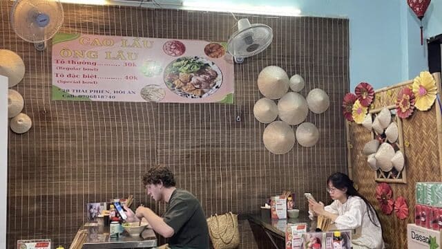 Two people eating at a small Vietnamese restaurant with bamboo decor and a menu sign on the wall.