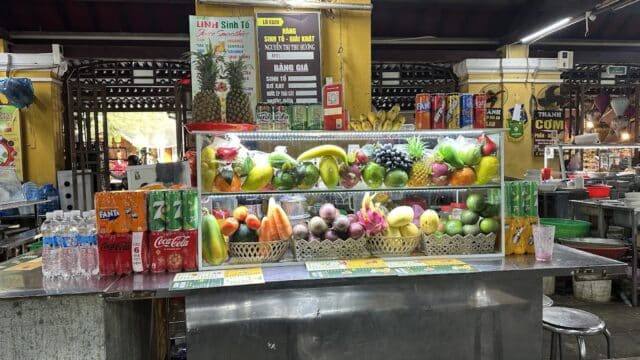 A colorful fruit and drink stand in Hoi An market, with fresh fruits, bottled water, and canned sodas on display.