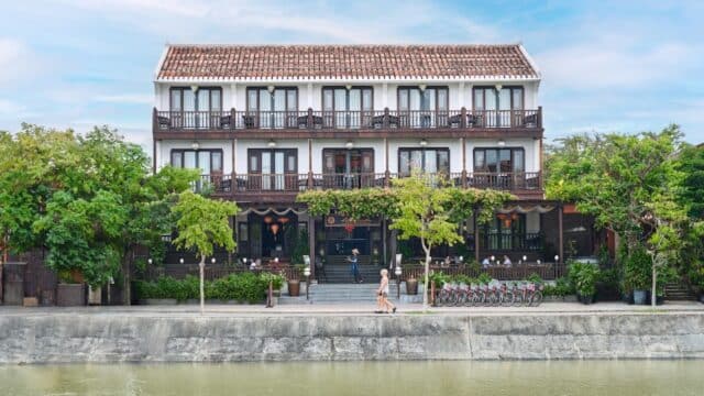 Three-story riverside boutique hotel with balconies, surrounded by trees, people, and bicycles—enjoy the charm of Little Hoi An.