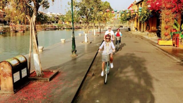 Three people with Local Buddy Tours ride bicycles along a riverside street lined with trees and lanterns on a sunny day.