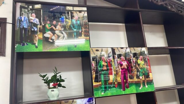 Framed photos of people in colorful outfits by a local tailor displayed on a wooden shelf with a potted plant below.