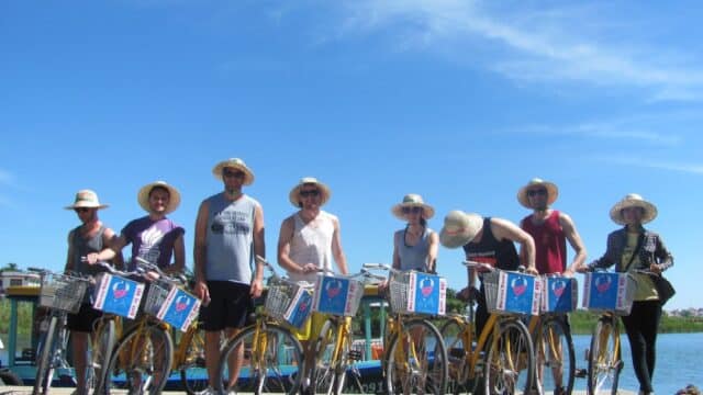 Eight people wearing hats pose with yellow bicycles by the water, sharing their love of life under a clear blue sky.