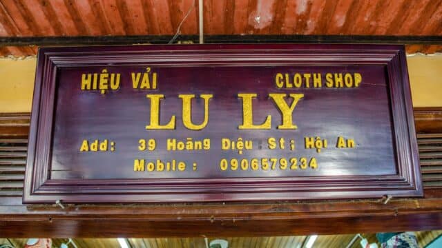 Wooden shop sign for LU LY Cloth Shop in Hoi An, Vietnam, featuring address and phone number.
