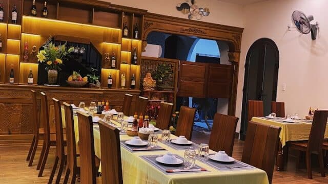 A warmly lit dining room at Madame Hạnh Restaurant, with wooden tables, chairs, and bottles displayed on decorated shelves.