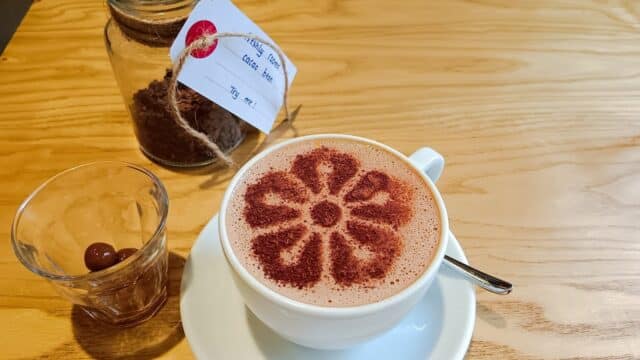 A Maison Marou cappuccino with cocoa flower art, a small glass, and a jar of cocoa powder on a wooden table.