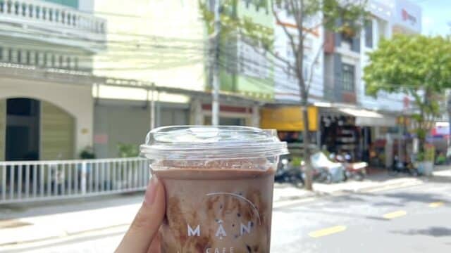 A hand holds an iced chocolate drink in a clear cup on a sunny street in Hoi An, with charming buildings in the background.