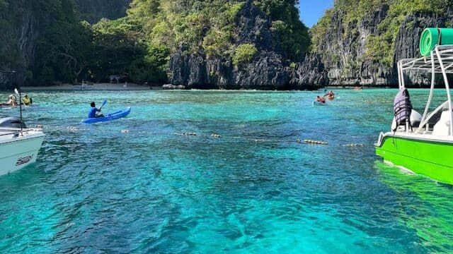 Clear turquoise water, kayakers, and boats surrounded by lush green cliffs under a bright blue sky.