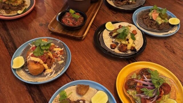 Assorted tacos from Masa Taqueria on colorful plates with lime wedges and drinks, served in a lively Mexican restaurant setting.