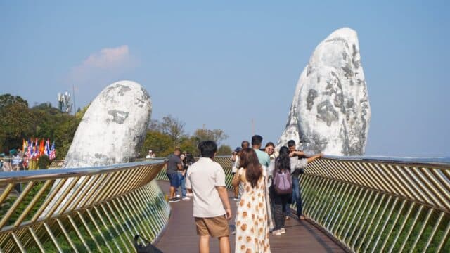 People walk on a bridge supported by giant stone hands under a clear blue sky.