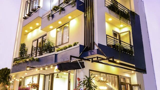 Three-story modern building with balconies and warm lights, surrounded by plants at dusk.