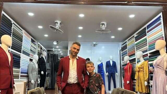 Man in a red suit and a boy pose inside a tailor shop, surrounded by mannequins and colorful fabric rolls.