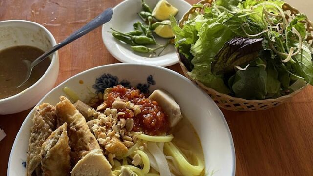 A bowl of Vietnamese noodles with tofu, sauce, herbs, and fresh greens inspired by the flavors of Hoi An on a wooden table.