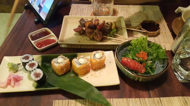 A table in Hoi An with sushi rolls, skewers, salad, dipping sauces, and a glass of water on placemats.