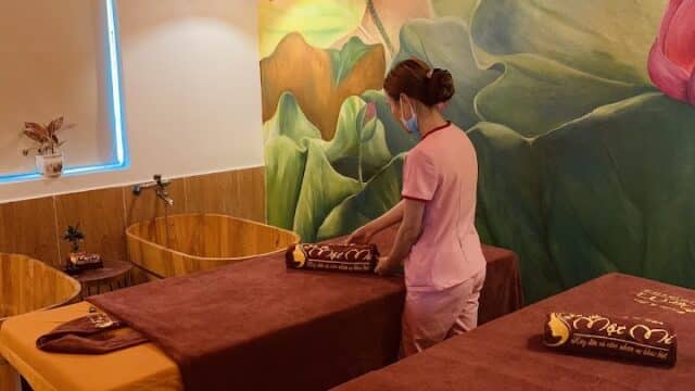 A person prepares massage tables in a Beauty Spa room at Một Mí Beauty with lotus flower wall art and warm lighting.
