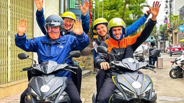 Four smiling people wearing helmets wave while sitting on two motorbikes on a city street near a spa.