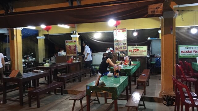 Outdoor restaurant, Mr. Sơn Restaurant features green tablecloths, wooden benches, and diners enjoying meals under bright lights at night.