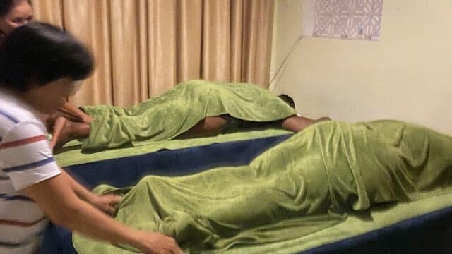Two people lying on massage tables covered with green sheets; someone adjusts a sheet on one table.