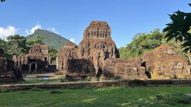 Ancient brick temple ruins near Hoi An, surrounded by greenery and trees, with a mountain backdrop under a clear blue sky.