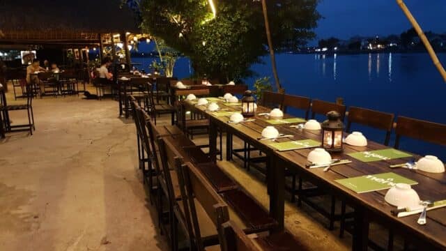 Nem Kitchen Riverside: Outdoor riverside restaurant at dusk with set tables, chairs, and lanterns, overlooking calm water.