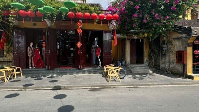Street view of a Hoi An building with red lanterns, green hanging decorations, and a flowering tree under a blue sky.
