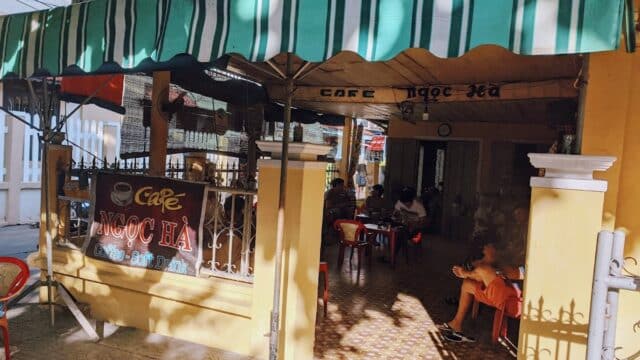 Outdoor view of a yellow Hoi An café with green-striped awning and people sitting inside and outside.
