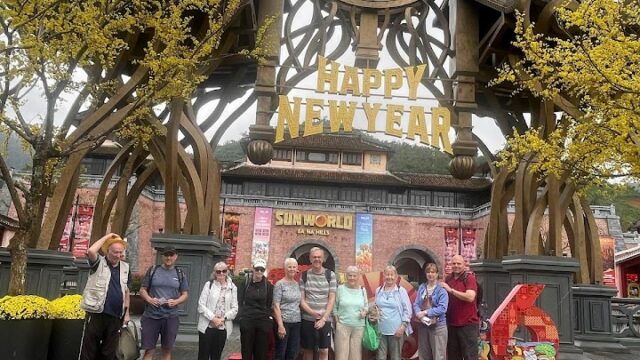 A group poses under a large Happy New Year sign at the Sun World entrance, surrounded by yellow trees on a Nguyen Tours adventure.