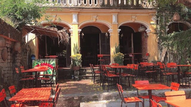 Outdoor cafe with red tables and chairs, shaded by trees, in front of a yellow colonial-style building in Hoi An.