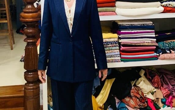 Smiling woman in a pinstripe suit stands barefoot in front of shelves stacked with colorful Hoi An fabric rolls.
