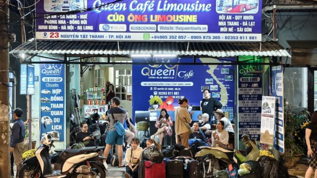 People with luggage wait outside a brightly lit Queen Cafe Limousine bus office in Hoi An at night, with scooters parked nearby.