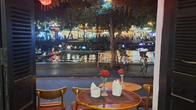 A cozy café table by open doors at Nhat Anh Restaurant, overlooking lantern-lit trees and a river at night.